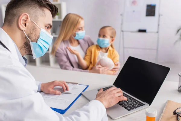 Pediatra em máscara médica usando laptop perto de pílulas e família na clínica — Fotografia de Stock