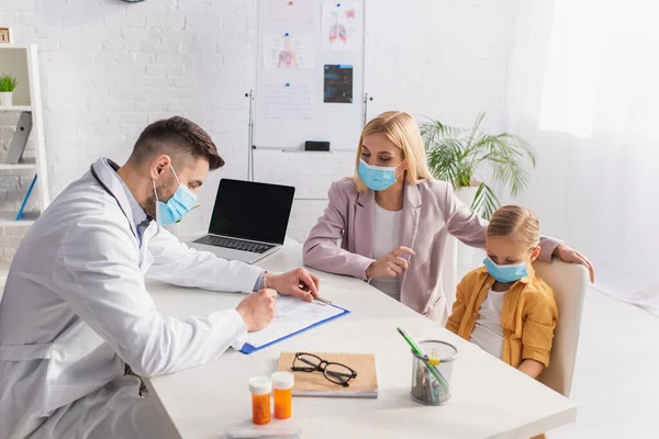Pediatrician in protective mask writing on clipboard near mother and patient — Stock Photo