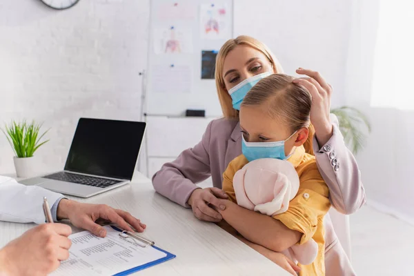 Мать смотрит на дочь в медицинской маске рядом с семейным врачом, пишущим на планшете — стоковое фото
