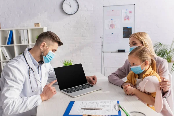 Arzt in medizinischer Maske zeigt auf Laptop in der Nähe von Mutter und Kind — Stockfoto