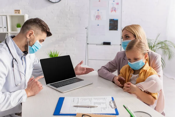 Médico señalando a la computadora portátil y la familia en máscaras médicas en la clínica - foto de stock