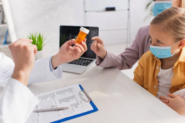Mujer señalando con el dedo cerca de médico de familia con pastillas e hija en máscara médica - foto de stock