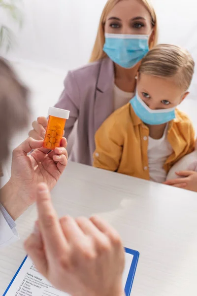 Pastillas en la mano de pediatra borrosa y madre con niño en máscaras médicas - foto de stock