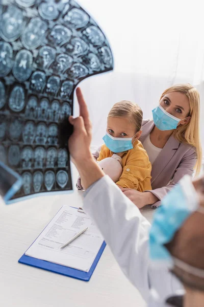 Мать и ребенок в медицинских масках смотрят на педиатра с МРТ на размытом переднем плане — стоковое фото
