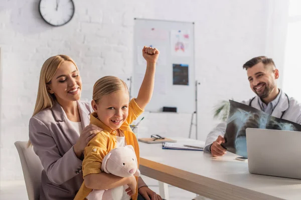 Lächelndes Kind zeigt Ja-Geste nahe Mutter und verschwommenem Arzt mit Fluorographie — Stockfoto