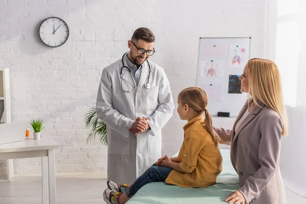 Smiling family doctor looking at kid on medical couch near mother — Stock Photo