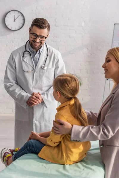 Positive pediatrician looking at girl on medical couch near mother — Stock Photo