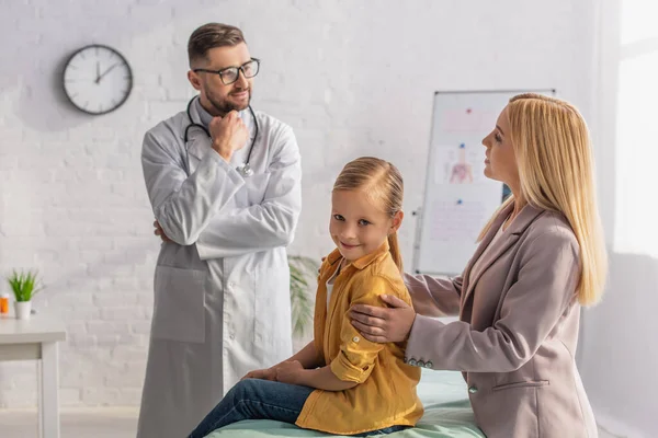 Lächelndes Kind blickt in Kamera nahe Mutter im Gespräch mit Kinderarzt — Stockfoto