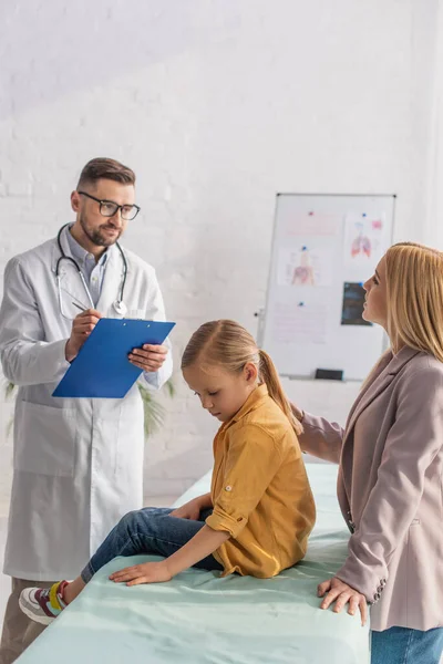 Pediatrician writing on clipboard near mother and sad kid in clinic — Stock Photo