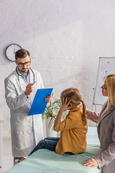Doctor with clipboard looking at sick child on medical couch near parent — Stock Photo