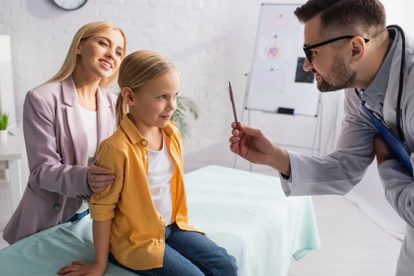 Pediatrician holding pen near kid and smiling mother in clinic — Stock Photo