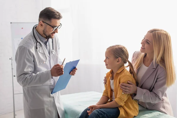 Médico con portapapeles y bolígrafo de pie cerca de la madre sonriente abrazando al niño en el sofá médico - foto de stock