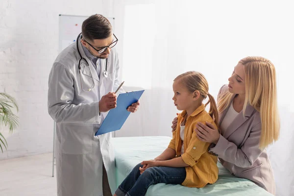 Doctor with pen and clipboard looking at girl on medical couch near parent — Stock Photo