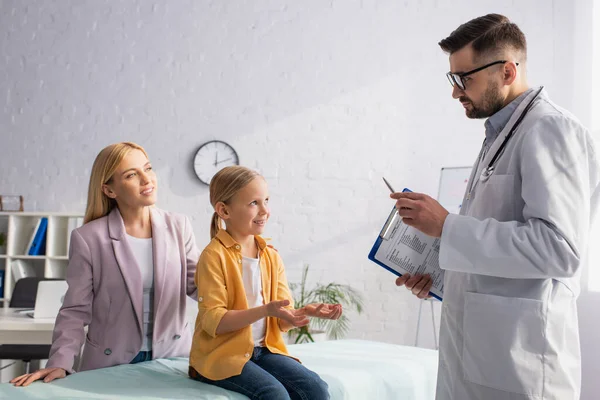 Enfant souriant parlant au pédiatre avec presse-papiers et mère à la clinique — Photo de stock