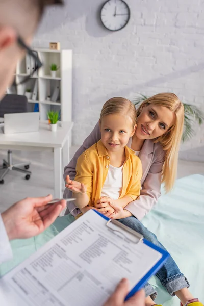 Kid talking to blurred pediatrician with blurred clipboard near smiling mother in hospital — Stock Photo