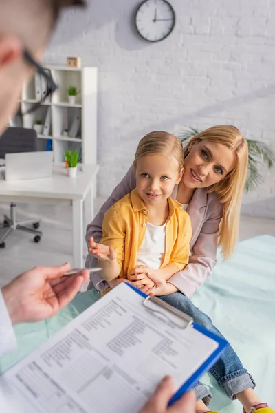 Kind spricht mit verschwommenem Kinderarzt mit verschwommenem Klemmbrett neben lächelnder Mutter im Krankenhaus — Stockfoto