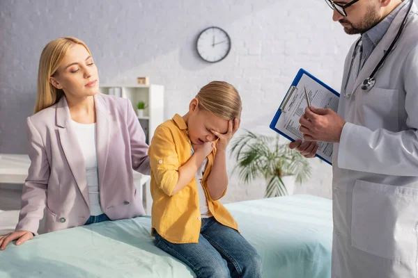 Verärgertes Mädchen sitzt auf medizinischer Couch in der Nähe von Arzt und Mutter — Stockfoto