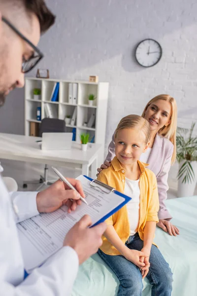 Smiling girl looking at doctor writing on clipboard near mother in clinic on blurred background — Stock Photo