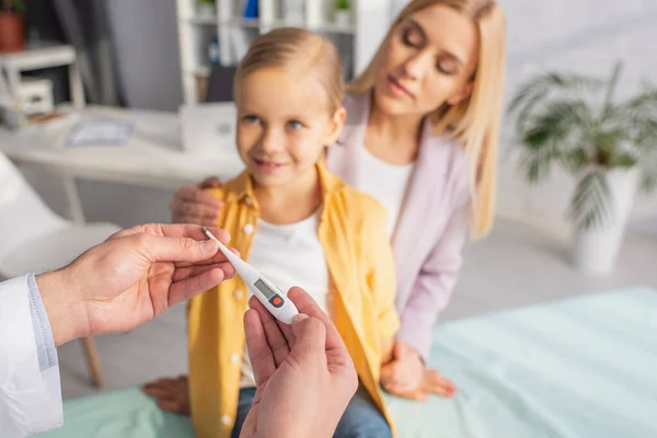 Arzt hält Thermometer in der Nähe von Mutter und Kind auf verschwommenem Hintergrund — Stockfoto