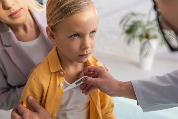 Médecin de famille tenant un thermomètre près d'un enfant et une mère floue — Photo de stock