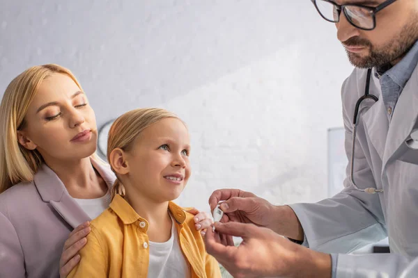 Doctor sosteniendo termómetro electrónico cerca de niño sonriente y padres en el hospital - foto de stock