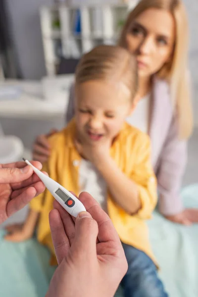 Médico sosteniendo termómetro cerca del niño enfermo y la madre sobre fondo borroso - foto de stock