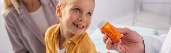 Medico in possesso di barattolo con pillole vicino bambino sorridente e madre offuscata, striscione — Foto stock
