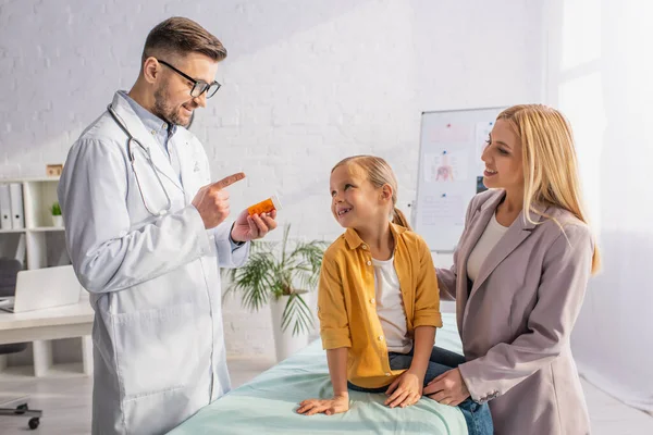 Усміхнений лікар тримає таблетки і вказує пальцем на матір і дитину на медичному дивані — стокове фото