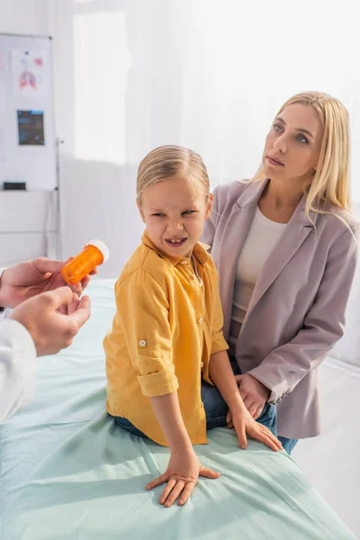 Pediatra sosteniendo pastillas cerca de niño disgustado en el sofá médico y la madre - foto de stock