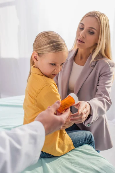 Pediatra borrosa sosteniendo frasco con pastillas cerca de la madre y paciente disgustado - foto de stock