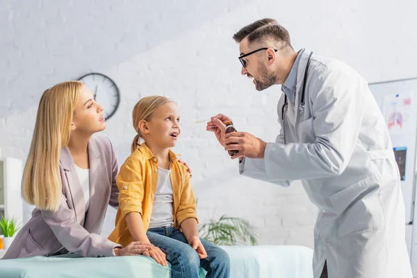 Arzt mit Sirup in der Nähe von Kind mit offenem Mund und Mutter in Klinik — Stockfoto