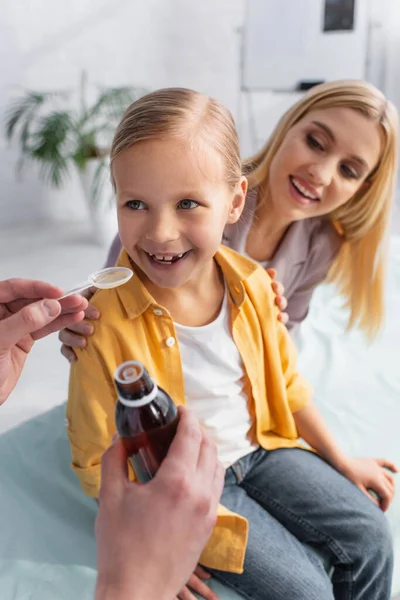 Médico sosteniendo cuchara de jarabe cerca de niño positivo y madre sonriente sobre fondo borroso - foto de stock