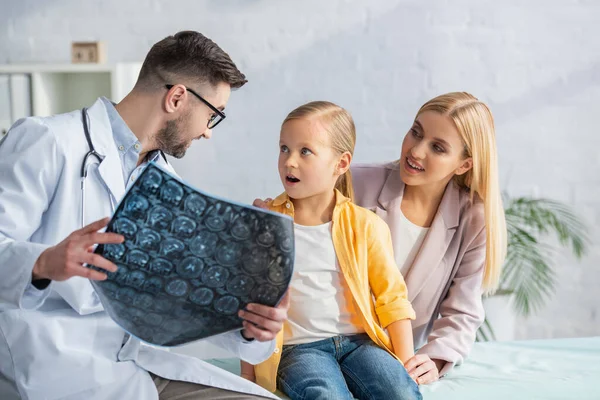 Pediatra sosteniendo borrosa resonancia magnética cerca de niño y madre asombrados - foto de stock