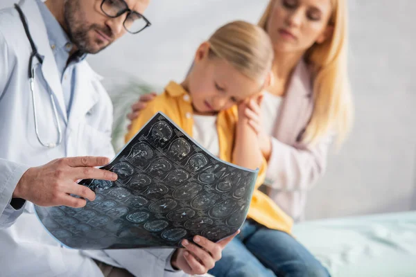 Mri scan in hands of blurred pediatrician near sad kid and woman in clinic — Stock Photo