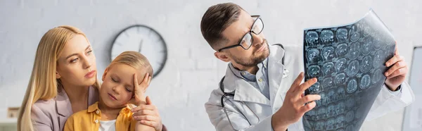 Pediatra em óculos olhando para a ressonância magnética perto da mãe e criança doente, banner — Fotografia de Stock