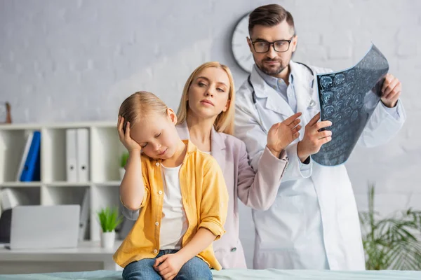 Miúdo mal-humorado a tocar na cabeça perto da mãe turva e pediatra com ressonância magnética no hospital — Fotografia de Stock