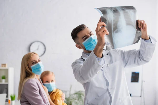 Pediatra en máscara médica mirando la fluorografía cerca de madre borrosa abrazando niño - foto de stock