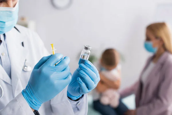 Médico em máscara médica segurando vacina perto de mãe e criança turva — Fotografia de Stock