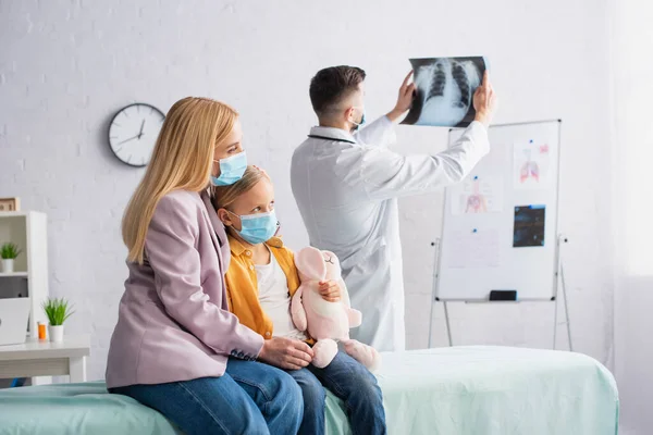 Жінка в медичній масці обіймає дитину на медичному дивані біля лікаря з флюорографією в клініці — стокове фото