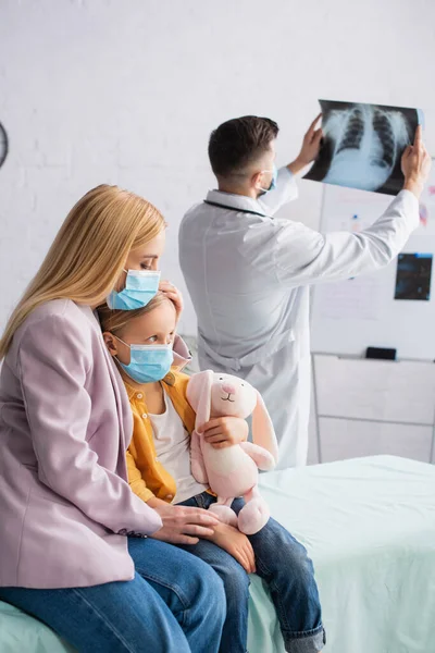 Mujer con máscara médica sentada cerca de un niño en un sofá médico y pediatra borroso con fluorografía - foto de stock