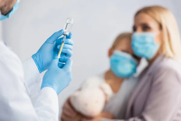 Врач в латексных перчатках собирает вакцину в шприце рядом с матерью и ребенком на размытом фоне — стоковое фото