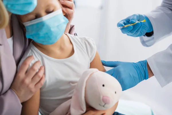 Doctor in latex gloves holding syringe near mother hugging kid in medical mask — Stock Photo