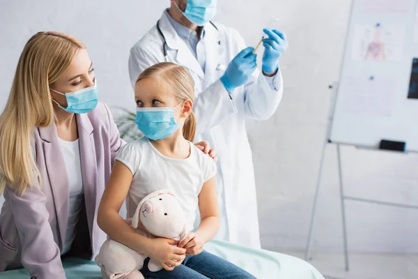 Mother in medical mask looking at daughter with soft toy near doctor with vaccine in hospital — Stock Photo
