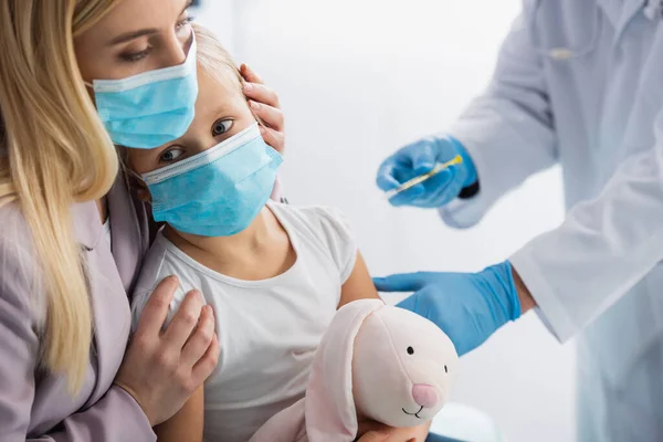 Mother hugging kid in medical mask near pediatrician with syringe on blurred background — Stock Photo