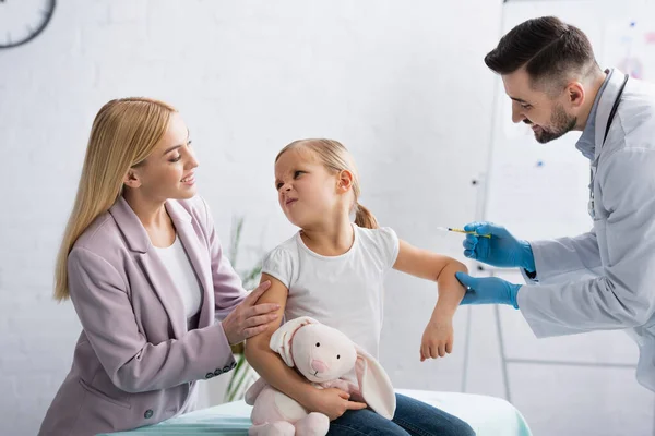 Kid looking at smiling mother near pediatrician with syringe — Stock Photo