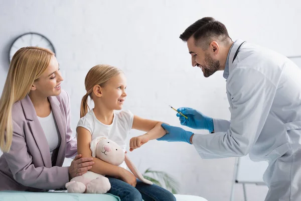 Happy kid looking at doctor with syringe near mother in hospital — Stock Photo