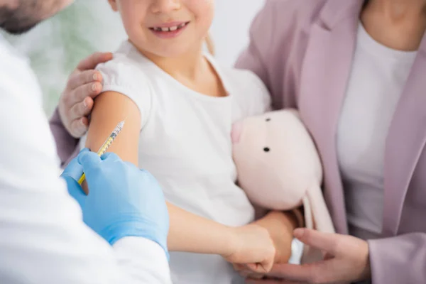 Cropped view of doctor holding syringe near smiling kid with soft toy and mother — Stock Photo