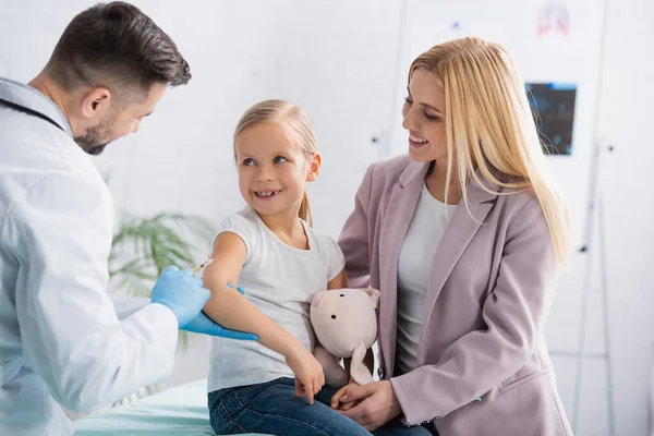 Smiling parent looking at kid near doctor with syringe during vaccination — Stock Photo