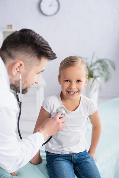 Pediatra borroso usando estetoscopio en niño alegre en la clínica - foto de stock