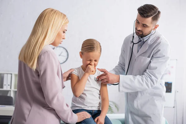 Niño enfermo estornudando cerca de los padres y pediatra con estetoscopio - foto de stock
